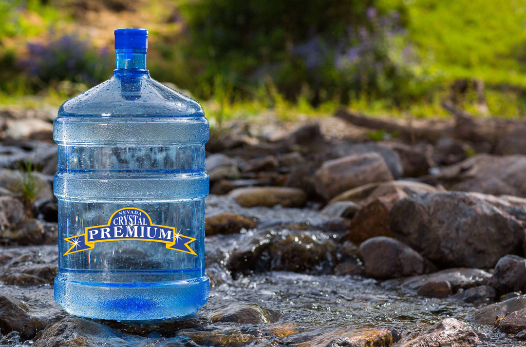 A large water jug labeled "Nevada Crystal Premium" stands on rocks in a flowing stream surrounded by greenery.
