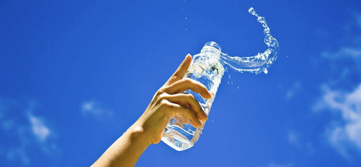 A hand holds a clear plastic bottle of alkaline water against a blue sky, with water splashing out, highlighting the health benefits of making the switch to alkaline water.