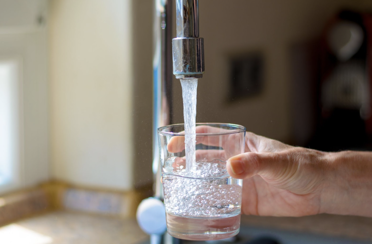 Bildschirmfoto 2025-12-07 um 15.46.39 A hand is holding a glass under a running kitchen faucet, filling it with tap water.