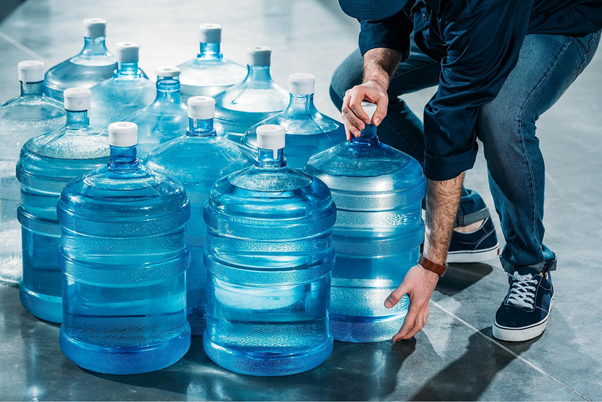 image1 A person in casual clothing lifts a large blue water jug among several similar jugs on a shiny floor, showcasing the convenience of home water delivery.
